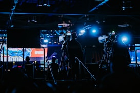 Silhouettes of camera operators and equipment set up in a dimly lit venue, illuminated by blue stage lights. A large screen displays colorful graphics in the background.