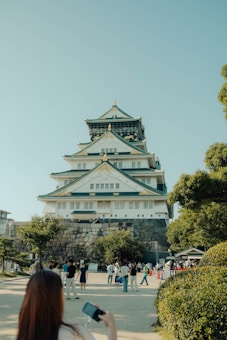 A majestic traditional-style castle with multiple tiers, surrounded by stone walls and lush greenery. A crowd of people is gathered in front, some taking photos. The sky is clear and blue, conveying a serene atmosphere.