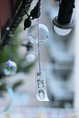 A delicate glass wind chime hangs from a string, with a blue-and-white paper strip attached. The chime is adorned with subtle floral patterns in muted colors. In the background, a soft-focus urban setting with other hanging objects and blurred lights.
