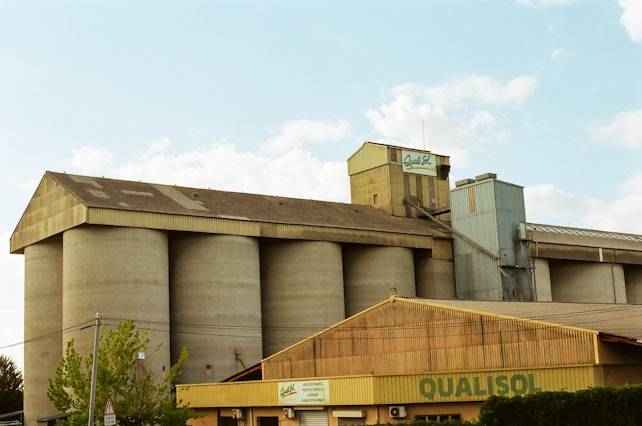 A large industrial building with multiple cylindrical silos and a sloped roof. The structure is constructed from concrete and metal, with 'QUALISOL' signage displayed prominently. A smaller building with a similar roof style is in the foreground, partially obstructed by trees and power lines. The sky above is partly cloudy.