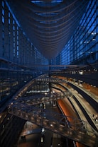 A modern architectural interior features a vast, sweeping structure with high ceilings and expansive glass walls. A central curved walkway and multiple levels are illuminated by soft lights, creating a sense of spaciousness and elegance. The design incorporates steel and glass elements, emphasizing a futuristic and sleek aesthetic.