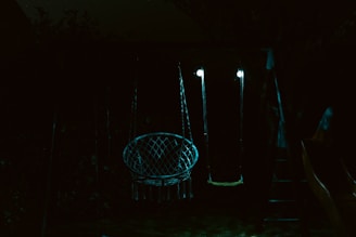 A dimly lit outdoor swing set featuring two swings, one with a net-like structure and the other a simple flat seat, both illuminated by faint lights. The surrounding environment is dark with hints of foliage in the background.
