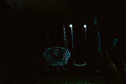 A dimly lit outdoor swing set featuring two swings, one with a net-like structure and the other a simple flat seat, both illuminated by faint lights. The surrounding environment is dark with hints of foliage in the background.