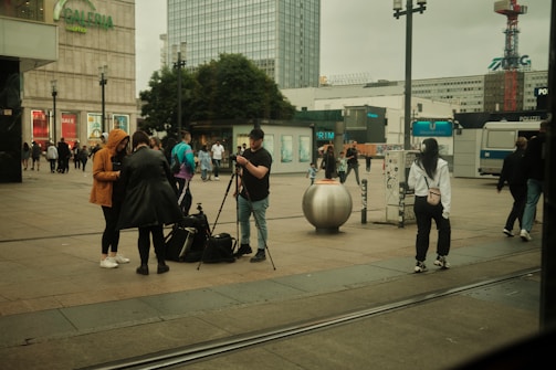 A busy urban street scene with people walking and interacting. A man stands with a camera on a tripod, engaging with others around him. An advertisement for a sale is visible on a large building labeled Galeria. Trees and a modern glass building form the background.