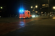An ambulance with blue lights flashing is driving through a dimly lit urban area at night. The road is wet, reflecting the streetlights, which illuminate the otherwise dark scene.