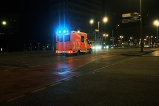 An ambulance with blue lights flashing is driving through a dimly lit urban area at night. The road is wet, reflecting the streetlights, which illuminate the otherwise dark scene.