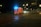 An ambulance with blue lights flashing is driving through a dimly lit urban area at night. The road is wet, reflecting the streetlights, which illuminate the otherwise dark scene.