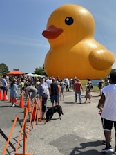 A large, inflatable yellow rubber duck dominates the scene, surrounded by a lively crowd enjoying an outdoor event. The duck features bright orange beak and black eyes, and people of various ages are gathered around, some with pets. The sky is clear and blue, adding to the vibrant atmosphere, with orange traffic cones and temporary barriers in place.