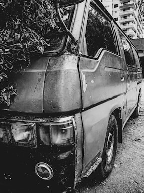 An old, worn-out van is parked on a rough, unpaved surface. The vehicle's exterior is weathered and rusted, with peeling paint. Foliage encroaches on one side, suggesting neglect and the passage of time. In the background, there are tall buildings, indicating an urban environment.