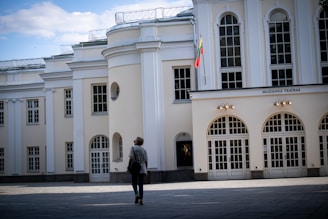 A person is walking towards a white classical building with large arched windows. The structure has an ornate, architectural style with detailed columns and symmetrical design. The flag of Lithuania is displayed near the entrance, indicating the building's national significance. The sky is partly cloudy, contributing to the serene atmosphere.
