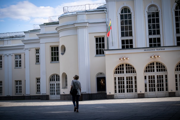 A person is walking towards a white classical building with large arched windows. The structure has an ornate, architectural style with detailed columns and symmetrical design. The flag of Lithuania is displayed near the entrance, indicating the building's national significance. The sky is partly cloudy, contributing to the serene atmosphere.