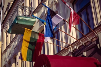 Several national flags are displayed on flagpoles attached to the side of a building with ornate architectural details. The flags include Lithuania, Latvia, and Estonia, indicating a representation of Baltic states. The building features decorative ironwork on a balcony and large windows.