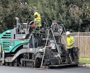 Two construction workers in high-visibility vests and hard hats are operating a large road paver machine. They are laying fresh asphalt on a road, with one worker standing on the machine and the other standing on the ground beside it. The scene is set outdoors with some trees and a wooden fence in the background.