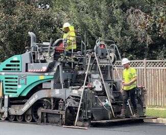 Two construction workers in high-visibility vests and hard hats are operating a large road paver machine. They are laying fresh asphalt on a road, with one worker standing on the machine and the other standing on the ground beside it. The scene is set outdoors with some trees and a wooden fence in the background.