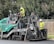 Two construction workers in high-visibility vests and hard hats are operating a large road paver machine. They are laying fresh asphalt on a road, with one worker standing on the machine and the other standing on the ground beside it. The scene is set outdoors with some trees and a wooden fence in the background.