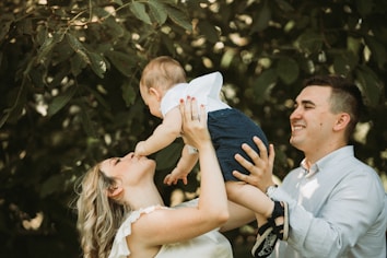 A smiling couple interacts with their baby outdoors. The mother lifts the child towards her face, and the father stands beside her, holding the baby as well. They are surrounded by foliage, suggesting a natural, serene environment.