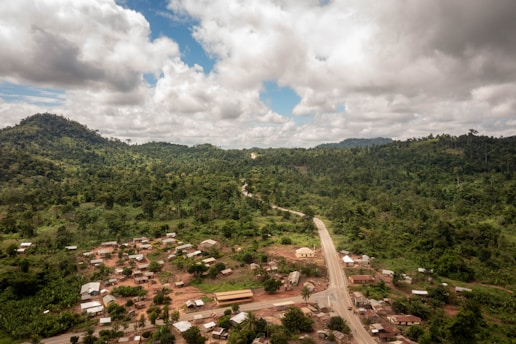 Aerial view of a rural landscape featuring a small village nestled amidst dense, lush green forest. A narrow road winds through the settlement, which consists of numerous small buildings with tin roofs. Rolling hills covered with thick vegetation are visible in the distance under a partly cloudy sky.