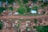 An aerial view of a small village or rural area with scattered houses, most of which have rust-colored roofs. A dirt road runs through the center, with a few vehicles visible. Surrounding the houses are areas of green vegetation, indicating grassy or possibly forested areas. The layout suggests an informal settlement with unevenly spaced buildings.