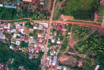 An aerial view of a small town with unpaved roads and various buildings. The landscape is a mix of greenery and residential structures. The roads intersect at different points, forming a network that connects the scattered houses. Some roofs are brightly colored, contrasting with the surrounding green areas and brown earth.