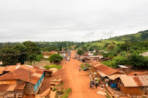 A small rural village with dirt roads and simple houses with tin roofs. There are people walking and a few motorbikes and vehicles on the road. The area is surrounded by lush green vegetation and hills in the background.