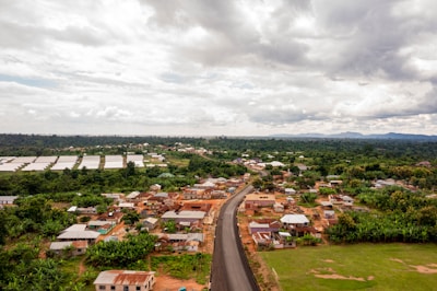 A rural landscape with a road stretching through a small settlement of houses with tin roofs. The area is surrounded by green vegetation and farmland. In the distance, there are lush forests and hills under a cloudy sky.