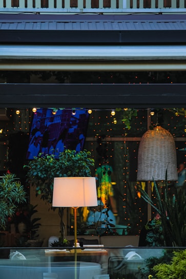 A cozy interior space with various elements including a lit floor lamp on a wooden table, surrounded by lush green plants. A large wicker pendant light hangs from the ceiling, and the room is decorated with strings of small, warm fairy lights. The ambiance is inviting, with a mixture of natural and artificial elements creating a warm and intimate atmosphere.