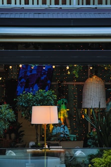 A cozy interior space with various elements including a lit floor lamp on a wooden table, surrounded by lush green plants. A large wicker pendant light hangs from the ceiling, and the room is decorated with strings of small, warm fairy lights. The ambiance is inviting, with a mixture of natural and artificial elements creating a warm and intimate atmosphere.
