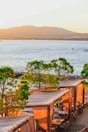 A picturesque coastal scene featuring a waterfront restaurant with wooden cabanas covered by beige drapes. The cabanas are nestled among green trees, and the backdrop includes a calm blue water body with small boats and distant mountains under a warm sunset.