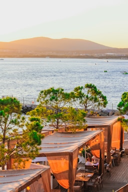A picturesque coastal scene featuring a waterfront restaurant with wooden cabanas covered by beige drapes. The cabanas are nestled among green trees, and the backdrop includes a calm blue water body with small boats and distant mountains under a warm sunset.