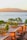 A picturesque coastal scene featuring a waterfront restaurant with wooden cabanas covered by beige drapes. The cabanas are nestled among green trees, and the backdrop includes a calm blue water body with small boats and distant mountains under a warm sunset.