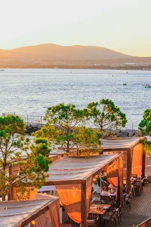 A picturesque coastal scene featuring a waterfront restaurant with wooden cabanas covered by beige drapes. The cabanas are nestled among green trees, and the backdrop includes a calm blue water body with small boats and distant mountains under a warm sunset.