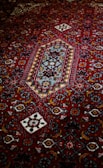 An intricately designed carpet featuring a rich red background with geometric and floral patterns in contrasting colors, including white, blue, and darker shades. The central motif is a diamond shape surrounded by a series of decorative elements.