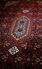 An intricately designed carpet featuring a rich red background with geometric and floral patterns in contrasting colors, including white, blue, and darker shades. The central motif is a diamond shape surrounded by a series of decorative elements.