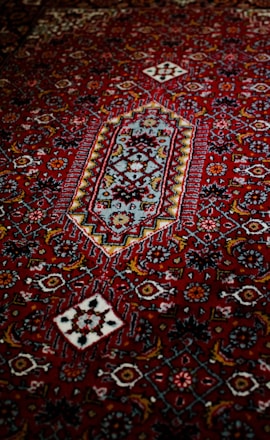 An intricately designed carpet featuring a rich red background with geometric and floral patterns in contrasting colors, including white, blue, and darker shades. The central motif is a diamond shape surrounded by a series of decorative elements.