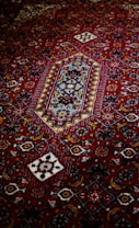 An intricately designed carpet featuring a rich red background with geometric and floral patterns in contrasting colors, including white, blue, and darker shades. The central motif is a diamond shape surrounded by a series of decorative elements.