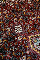 Intricate geometric patterns in vibrant colors cover the surface of a richly textured fabric. The composition includes repeating motifs in shades of red, blue, yellow, and white, creating an elaborate tapestry with floral and angular designs.