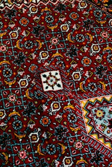 Intricate geometric patterns in vibrant colors cover the surface of a richly textured fabric. The composition includes repeating motifs in shades of red, blue, yellow, and white, creating an elaborate tapestry with floral and angular designs.