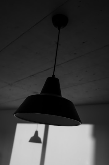 A black and white image featuring a ceiling-mounted pendant lamp casting a distinct shadow on the wall. The setting conveys a minimalist, industrial interior with clean lines and a smooth surface on the ceiling.