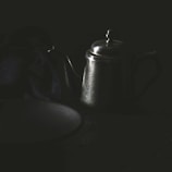 A dark, moody still life featuring a metallic teapot with a long spout and a tidy lid. The teapot is placed on a dimly lit surface, accompanied by a piece of dark fabric that drapes elegantly beside it. The lighting casts soft reflections off the teapot, adding a mysterious ambiance to the scene.