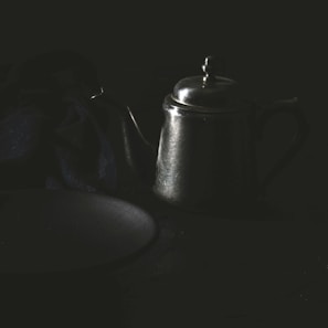 A dark, moody still life featuring a metallic teapot with a long spout and a tidy lid. The teapot is placed on a dimly lit surface, accompanied by a piece of dark fabric that drapes elegantly beside it. The lighting casts soft reflections off the teapot, adding a mysterious ambiance to the scene.