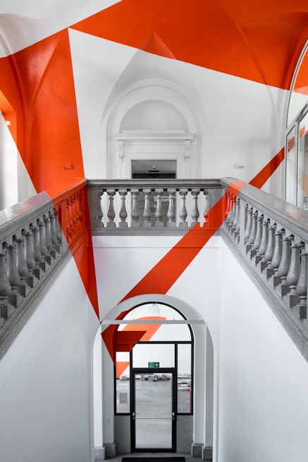 A staircase with a modern art installation featuring bold, angular orange shapes painted across the walls and ceiling. The architecture of the space includes classical elements such as decorative stone balustrades and high arched ceilings. The contrast between the traditional setting and the contemporary art creates a striking visual impact.