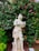 A classical stone statue of a person playing a musical instrument is set against a backdrop of lush green foliage and vibrant pink flowers. The figure appears to be wearing period clothing with intricate details. The surrounding garden is well-maintained, with various shades of green and pink adding to the visual richness.