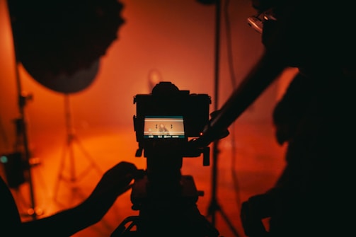 A camera setup is visible in a dimly lit room with a warm, orange tint. The camera is focused on a screen displaying a blurred image, while a person adjusts the equipment. The background includes shadows and professional lighting equipment.