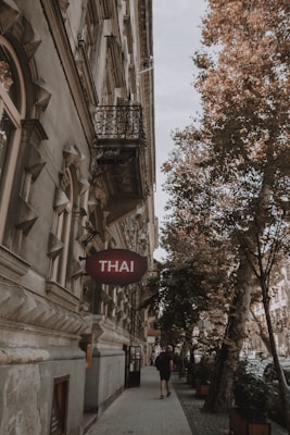 A street scene featuring a historic building with ornate architectural details and a sign reading 'Thai' indicating a restaurant. The sidewalk is lined with trees, and a person walks along the pavement, creating an inviting and quaint urban atmosphere.