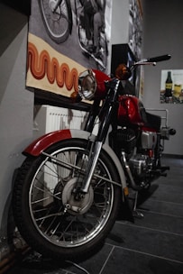 A vintage motorcycle is parked indoors near a wall, partially obscured by shadow. The wall is decorated with retro-style artwork and black-and-white photographs, adding a nostalgic feel to the setting. The motorcycle features a red fuel tank and chrome handlebars, reflecting a classic design.