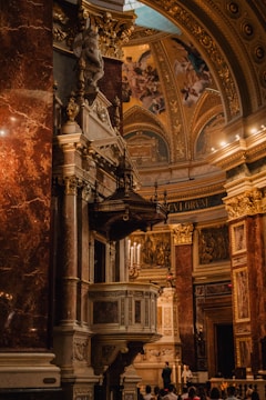 A richly decorated church interior featuring ornate architectural elements and detailed frescoes. Marble columns and intricate gold-tinted moldings accentuate the space. A pulpit is visible in the foreground, adorned with candles. The backdrop includes religious murals and arches, creating an atmosphere of historical grandeur.