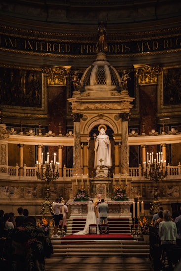 An ornate church interior with a couple at the altar during a wedding ceremony. The altar is adorned with candle stands and floral arrangements, and a large statue is prominently displayed above. The scene is illuminated by warm lighting, enhancing the grandeur of the surroundings.