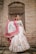 A woman stands elegantly against a rustic brick wall, wearing a traditional Indian lehenga adorned with intricate patterns. The outfit features a combination of white and pink with detailed embroidery. She accessorizes with traditional jewelry, including earrings and a headpiece, and her hair is styled neatly.