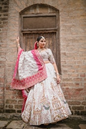 A woman stands elegantly against a rustic brick wall, wearing a traditional Indian lehenga adorned with intricate patterns. The outfit features a combination of white and pink with detailed embroidery. She accessorizes with traditional jewelry, including earrings and a headpiece, and her hair is styled neatly.