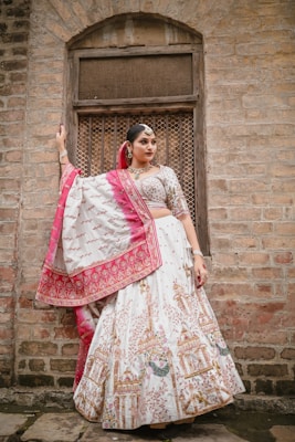 A woman stands elegantly against a rustic brick wall, wearing a traditional Indian lehenga adorned with intricate patterns. The outfit features a combination of white and pink with detailed embroidery. She accessorizes with traditional jewelry, including earrings and a headpiece, and her hair is styled neatly.